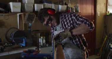 Beyaz erkek bıçak imalatçısı, zımpara makinesi kullanarak gözlük ve kulaklık takıyor. Bağımsız küçük işletmeci iş başında.