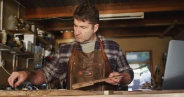 Kafkasyalı erkek bıçak üreticisi atölyede elinde bir aletle notlar tutuyor. Bağımsız küçük işletmeci iş başında.