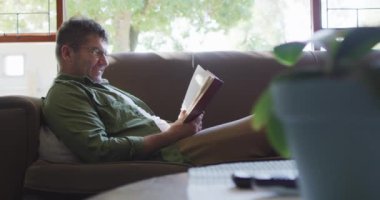 Beyaz adam kanepede oturuyor, evde kitap okuyor. ev hayatı ve boş zaman kavramı.