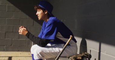 Nervous mixed race female baseball player, sitting on bench in sun waiting with baseball ball. female baseball team, sports training and game tactics.
