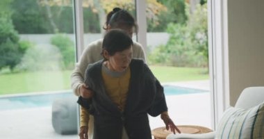 Yaşlı Asyalı annenin oturma odasında yetişkin bir kız tarafından sandalyeye oturtulması. Emeklilik hayatı, mutlu aile birlikte evde vakit geçirmek..