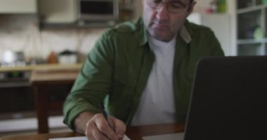 Beyaz adam masada oturuyor, dizüstü bilgisayar kullanıyor ve evde notlar alıyor. ev hayatı ve boş zaman kavramı.