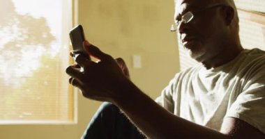 Düşünceli Afrikalı Amerikalı son sınıf öğrencisi evinde oturmuş akıllı telefon kullanıyor. Emeklilik hayatı, evde yalnız zaman geçirmek..