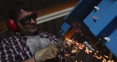 Kafkas erkek bıçak imalatçısı atölyede gözlük ve kulaklık takıyor, bileyici kullanıyor. Bağımsız küçük işletmeci iş başında.