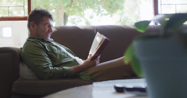 ソファに座って本を家で読んでいる白人男性 国内生活と余暇時間の概念 — ストック動画