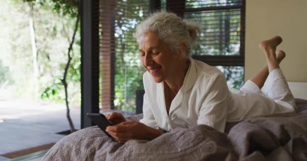 高级混血女子躺在床上 使用智能手机 面带微笑 退休和老年生活方式 独自呆在家里 — 图库视频影像