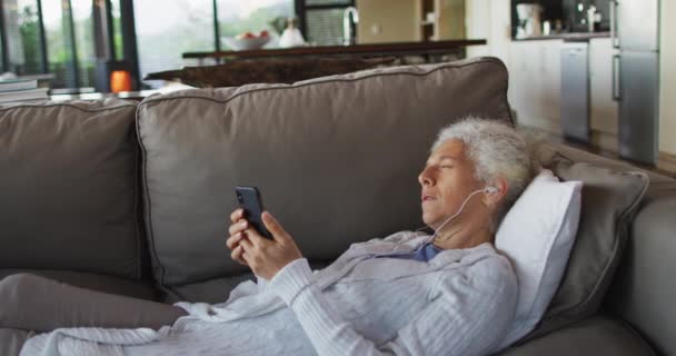 シニア混合レースの女性ソファにイヤホンを着用し スマートフォンを使用して横に 退職や高齢者のライフスタイル家で一人で過ごす — ストック動画
