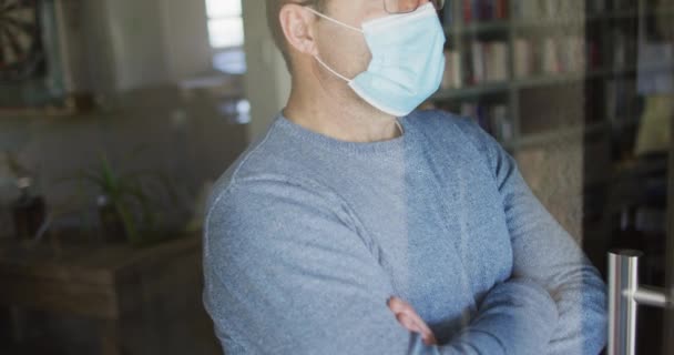 白人男性の顔のマスクを着て 窓から見ている コロナウイルスの流行期に家にいると — ストック動画