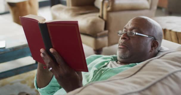 Χαρούμενος Αφροαμερικάνος Ηλικιωμένος Που Χαλαρώνει Ξαπλωμένος Στον Καναπέ Διαβάζοντας Ένα — Αρχείο Βίντεο
