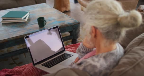 ラップトップを使用してソファに座っているシニア混合レースの女性 退職や高齢者のライフスタイル家で一人で過ごす — ストック動画