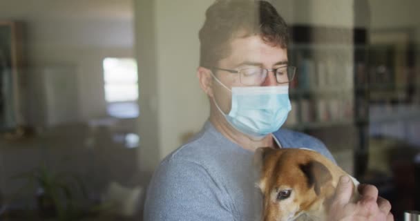 顔のマスクをした白人男性が窓に立って犬を撫でている コロナウイルスの流行期に家にいると — ストック動画
