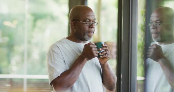 富有洞察力的非洲裔美国老人站在那里 喝着一大杯咖啡 望着窗外 退休生活方式 独自呆在家里 — 图库视频影像