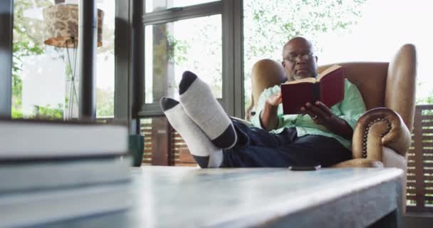 Счастливый Афроамериканец Пожилой Мужчина Расслабляется Кресле Поднятыми Ногами Читает Книгу — стоковое видео