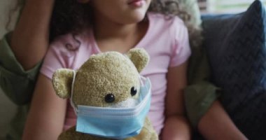 Karışık ırk anne-kız kucaklaşması, yüz maskeli oyuncak ayı tutan kız. covid 19 salgını sırasında aile ve aile eğlence zamanı konsepti.
