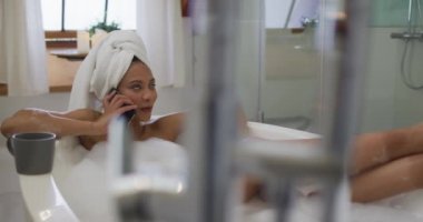 Karışık ırktan bir kadın banyo yapıyor ve akıllı telefondan konuşuyor. ev hayatı, kaliteli boş zaman geçirmek evde dinlenmek..