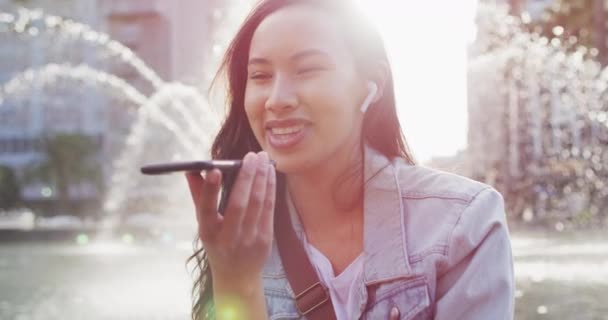 Asiatisk Kvinna Sitter Bredvid Fontänen Bär Hörlurar Och Talar Smartphone — Stockvideo