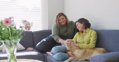 Kafkasyalı lezbiyen çift gülümsüyor ve köpekle kanepede oturuyor. Ev hayatı, evde dinlenerek boş zaman geçirmek..