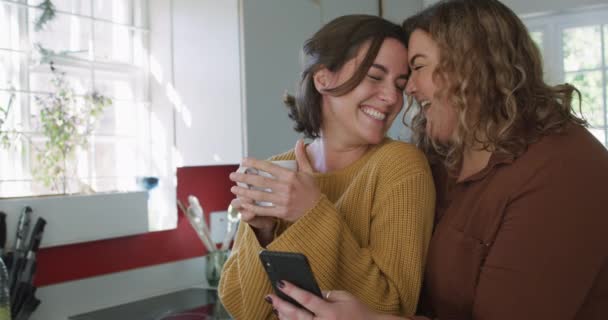 キッチンでスマホを抱きかかえ利用している幸せな白人レズビアンカップル 国内生活と余暇 — ストック動画