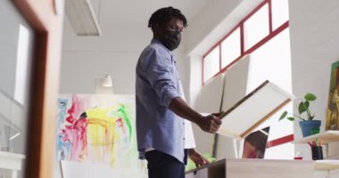 Yüz maskesi takan Afrika kökenli Amerikalı bir erkek sanatçı bir tablo tutuyor ve sanat stüdyosunda dizüstü bilgisayar kullanıyor. sanat, hobi ve yaratıcı meslek kavramı