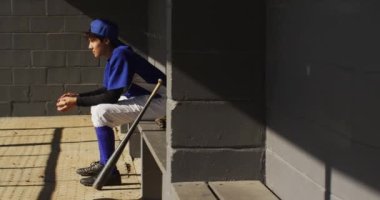 Sinirli melez bayan beyzbolcu, güneşin altında bankta oturmuş beyzbolla bekliyor. Kadın beyzbol takımı, spor antrenmanı ve oyun taktikleri.