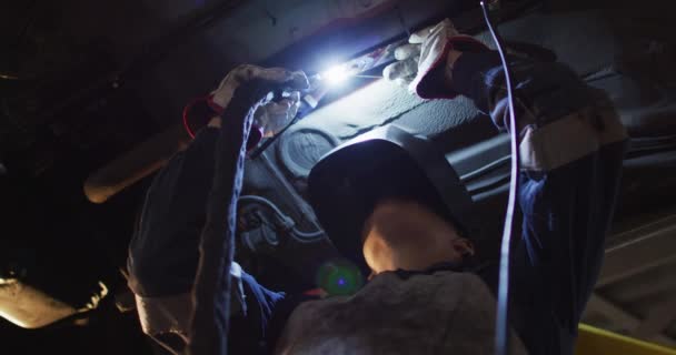 車のサービスステーションで車の下に溶接ヘルメット溶接を身に着けている女性整備士 自動車修理サービス — ストック動画