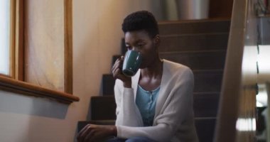 Düşünceli Afro-Amerikan çekici kadın merdivenlerde oturuyor ve kahve içiyor. Boş zaman, refah ve evde dinlenme..