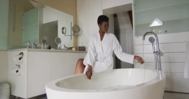 Beyaz bornozlu, gülümseyen Afro-Amerikan çekici kadın küvette oturmuş, banyoyu hazırlıyor. güzellik, şımartma, spa ve refah konsepti.