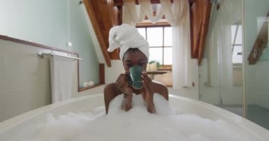Afro-Amerikan çekici kadın köpük banyosunda dinleniyor ve banyoda kahve içiyor. güzellik, şımartma, spa ve refah konsepti.