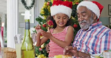 Mutlu Afro-Amerikan büyükbabası ve torunu Noel Baba şapkası takıp masada kutlama yapıyor. Aile Noel 'i ve şenliği evde birlikte kutluyoruz..