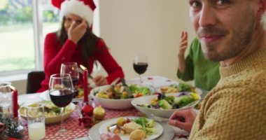 Noel yemeği sırasında kameraya bakan mutlu beyaz adam Noel Baba şapkası takıyor. Aile Noel 'i ve şenliği evde birlikte kutluyoruz..