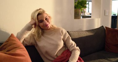 Düşünen kadın gri koltukta meraklı hissediyor, kameraya bakıyor, kırmızı atışı ayarlıyor, gülüyor. Düşünceli, rahat, rahat, yaşam tarzı, minimalist, ev, duygusal