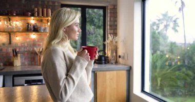 Mutfağa giriyor, elinde kırmızı kupa olan bir kadın pencereden dışarı bakıyor, sessiz bir yansıma arıyor. Huzur, evcil, rahat, huzur, yaşam tarzı, çağdaş, sıcak