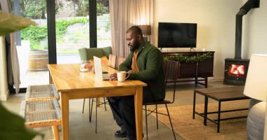 Yemek masasında dizüstü bilgisayarda yazı yazan Afro-Amerikalı bir adam. Kollarını gerdiğini hissediyor. Kopya uzayı. Teknoloji, çalışma alanı, rahatlama, ev, huzur, rahat, modern