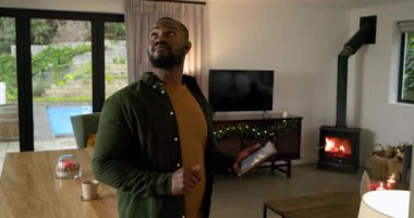 Afro-Amerikalı bir adam yemek masasında tablet tıkırdatıyor, tarama odasını tarıyor ve ev kontrollerini ayarlıyor. Teknoloji, akıllı ev, yaşam tarzı, çağdaş, rahatlama, iç mekan, otomasyon