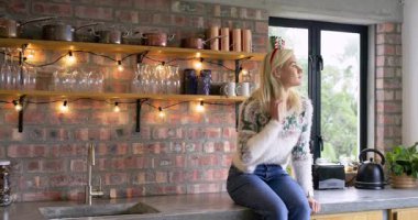 Kadın mutfak tezgahında oturmuş kameraya bakıyor. Saç bandıyla gülüyor, fotokopi çekiyor. Konforlu, rahat, rahat, yaşam tarzı, kırsal, sıcak, neşeli