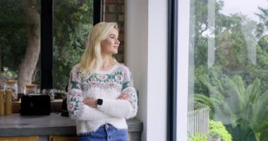 Mutfak tezgahına yaslanan, camdan bahçeye bakan, gülümseyen ve başını sallayan bir kadın. Düşünceli, rahat, rahat, modern, iç mekan, huzur, yaşam tarzı