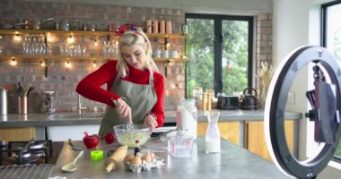 Mutfak adasında hamuru çırpan kadın akıllı telefon kamerasına bakıyor sonra da vanilya özü ekliyor. Yemek pişirme, aşçılık, yemek hazırlama, yaşam tarzı, ev mutfağı, organik