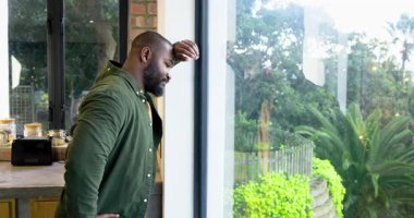Afro-Amerikalı bir adam mutfakta cam kapıya karşı durup, rahatlamak için boynunu esnetiyor. Yansıma, rahatlama, farkındalık, yeşillik, sükunet, sağIık, ev