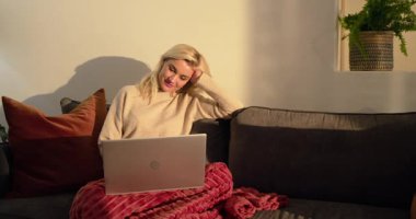 Yetişkin bir kadın kanepede dizüstü bilgisayarda dinleniyor. Kırmızı battaniyeli, yan yan bakan bir gülümsemeyle. Konfor, rahatlama, ev ofisi, konfor, rahatlık, ortam, modern