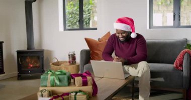 Afro-Amerikalı adam dizüstü bilgisayarını açıp oturma odasında Noel Baba şapkası takarak aileyi karşılıyor. Kutlama, sıcaklık, konfor, ev, dijital iletişim, tatil