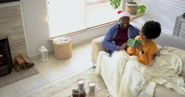 Afro-Amerikalı çift, battaniyenin altında evdeki kanepede gülüşlerini takiben tatil hediyelerini değiş tokuş ediyorlar. Samimiyet, samimiyet, kutlama, sıcaklık, şenlik, rahatlık, sevgi