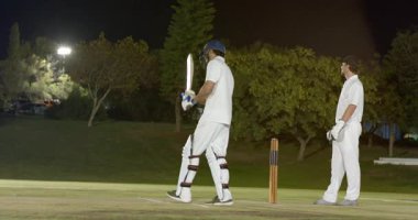 Bowlingci koşarken, Hintli vurucu sopayı vuruyor, eldivenleri ayarlıyor ve projektörlerin altında araba sürüyor. Kriket, gece, spor, yarışma, atletizm, performans, stadyum