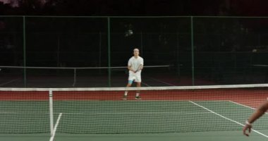 Sarı top servisi Asyalı bir adam ve partneri alacakaranlık kortunda voleybol oynayarak zaferi kutluyor. Spor, rekabet, takım çalışması, açık hava, dinamik, zafer, etkinlik kıyafeti