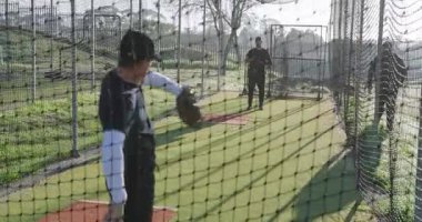 Koç ve stajyer hoplayan top el altından atılıyor ve vuruş kafesinde eldivenle yakalıyor. Spor eğitimi, beyzbol, antrenman, antrenman, atletizm, açık hava, yetenek geliştirme