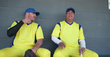 Ellerinde eldiven ve sopa olan takım arkadaşları yedek kulübesinde sohbet edip kahkahalarını paylaşıyorlar. Sporcular, takım çalışması, sportmenlik, dostluk, günlük, açık hava, rekabetçi