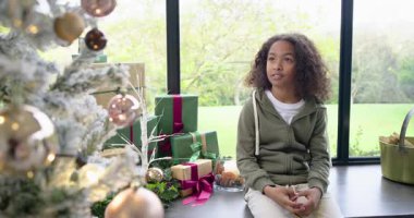 Çocuk pencere kenarındaki bankta oturuyor, ağaç süslerini fark ediyor ve hediyelere gülümserken bakışlarını değiştiriyor. Şen, neşeli, rahat, tatil, kış, kutlama, dekor