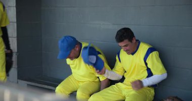 Paylaşılan şakalardan sonra gülen erkek beyzbol takımından arkadaşlar yedek kulübesinde kep ve rahatlatıcı takım arkadaşları. Atletizm, yoldaşlık, takım çalışması, rahatlama, gündelik, arkadaşlık, spor