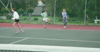 Top tepeye fırlatılıyor, bayan antrenör ve öğrenciler tenis kortunda forehand shot atıyorlar. Atletik, koçluk, eğitim, spor, açık hava, takım çalışması, odaklanma