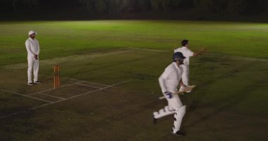 Çeşitli kriket oyuncuları sahada, bowlingde koşuyor, vurucular koşuyor, saha oyuncusu geri dönüyor. Spor, takım çalışması, atletik, yarışma, odak, açık hava, akşam