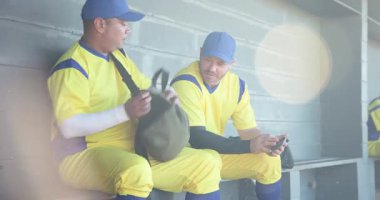 Yedek kulübesine giren çeşitli erkek beyzbol oyuncuları akıllı telefon özetlerini inceliyor, spor çantalarını açıyor. Sporcular, takım çalışması, eğlence, sportmenlik, günlük, açık hava, dostluk.